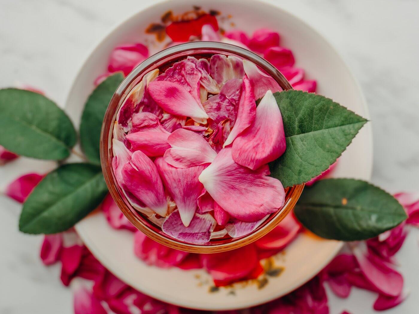 Glass bowl filled with pink rose petals and green leaves on a white plate