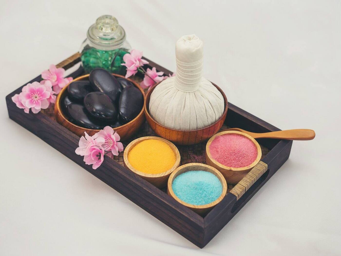 Wooden spa tray with smooth black stones, a herbal compress, bowls of colored bath salts, and pink flowers